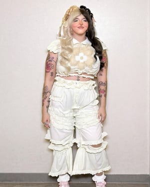 A woman, with puffy sleeves, off white puffy crop top and matching pants with holes, front light, white wallpaper background, cinematic, colorful tattoos, melanie martinez, full body, body glitter, full body standing infront of white wall, 4k, ultra realistic, photography, white lighting, pale, pink dress, light skin, white lighting, thin, curtain bangs, split hair blonde and black, chubby, smile, rosy cheeks, split hair pink and black, surreal, 8k, high quality, cinimatic, best quality, long pants, solid white background, long baggy off white pants, solid white background, very light pale skin tinted clothing, very ruffled shirt and pants, happy, smile, smile lines, real life, nose piercing, yellow tinted outfit, matte textured clothes, very baggy pants, oversized pants, happy, smile lines, smile, flower shape in the center of shirt, cinamatic, legs closed, hands on hips