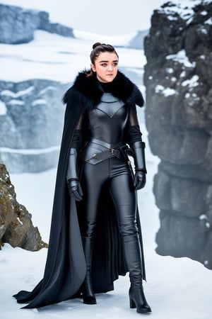 Maisie Williams stands tall in a winter wonderland setting, her long hair pulled back into a sleek bun. She wears a black leather armor set, with the open-ended cloak flowing behind her like a dark waterfall. A fur-lined collar adds warmth to her rugged ensemble as she gazes out into the snow-covered landscape.