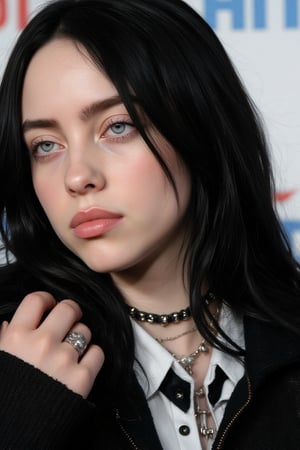 A close-up shot of Billie Eilish with long dark black hair and blue eyes. She is wearing a black jacket, a white collared shirt, a black tie, and a silver ring on her left hand. Her left hand is draped over her right shoulder. The backdrop is white with blue letters that read "HIT" and "HARD" and "SOFT" in red letters.