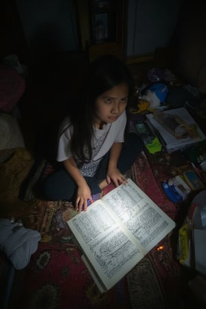 A high-angle close-up captures the distressed expression of a young Malay girl lit by a flashlight, her face contorted in frustration as she pores over a textbook. The room around her is chaotic, with scattered books and stationery adding to the sense of cluttered anxiety. Messy clothes and bedding cover the floor, amplifying the feeling of overwhelm.