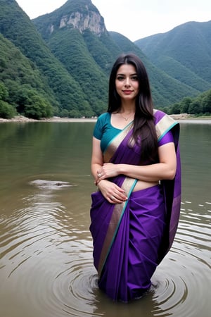 A woman with long, dark hair wearing a teal and purple saree standing in shallow water against a mountainous backdrop, with a relaxed yet stylish appearance.