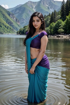 A woman with long, dark hair wearing a teal and purple saree standing in shallow water against a mountainous backdrop, with a relaxed yet stylish appearance.