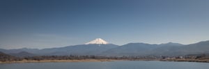 The fuji mountain captured at eye-level on a vibrant day, a majestic castle stands atop a mountain, adorned with a red roof. The castle is situated on a rocky cliff, adding a touch of nature to the scene. To the right of the castle, a stream of water is flowing, creating a peaceful and serene landscape. The sky is a deep blue, dotted with a few white fluffy clouds, adding depth to the composition.