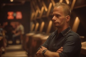 This photograph captures a light-skinned man with short, sandy blond hair and a slight stubble, standing with arms crossed in a dimly lit bar or club. He wears a black, collared shirt and a silver chain necklace, with a black wristwatch on his left wrist. The background is blurred, showing indistinct figures and warm, ambient lighting. The man exudes a confident, casual demeanor.,ATTA,#atta,#strsm