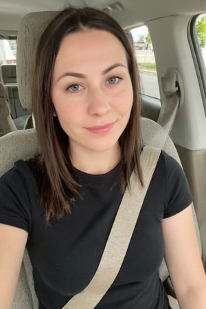 This is a photograph of a 30yo russian woman seated in the front passenger seat of a car. She has long, straight, dark brown hair that cascades over her shoulders, and she is wearing subtle makeup with a light pink lipstick. She has a fair complexion and is smiling gently at the camera. Her outfit consists of a simple, fitted black t-shirt. The car interior is visible, featuring gray fabric seats with visible stitching and a beige seatbelt across her body. The background outside the car window is blurred, showing hints of greenery, indicating the car is parked or moving slowly. The lighting is natural, suggesting daylight.