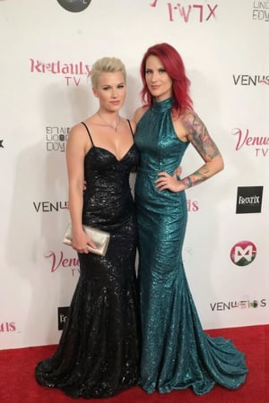 The image is a photograph taken at a formal event. It features two women standing closely together on a red carpet. The woman on the left has short, platinum blonde hair styled in a voluminous updo. She is wearing a dark, floor-length gown with thin straps and a fitted bodice adorned with sparkling sequins. She holds a silver clutch in her left hand and has a delicate necklace around her neck. The woman on the right has vibrant, bright red hair that cascades down her shoulders. She is dressed in a teal, floor-length gown with a high neckline and sleeveless design, featuring a shimmering texture that reflects the light. Her right arm is adorned with a colorful tattoo sleeve. Both women are smiling at the camera. The background is a white step-and-repeat banner with logos and text from sponsors such as "Reality TV," "Venus," "Beate Uhse," and "EroTIX." The lighting is bright, highlighting the details of their attire and the sparkle of their dresses. The overall atmosphere suggests a glamorous and celebratory event.