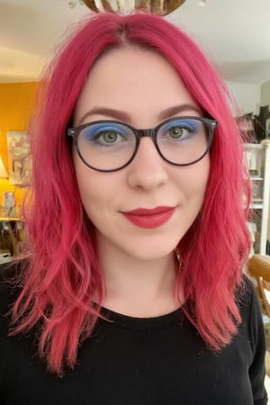 This is a close-up photograph of a 20yo western woman with striking features. She has vibrant, wavy pink hair cascading past her shoulders and wears black-rimmed glasses. Her skin is fair. Her eyes are green with bold blue eyeshadow, and she has a bright red lipstick on her full lips, which are slightly parted in a cheerful smile. She is wearing a black top. The background is slightly blurred but appears to be indoors, possibly a living room with warm lighting, wooden furniture, and some hanging decorations. The overall atmosphere is casual and vibrant, emphasizing the subject's colorful and expressive appearance.