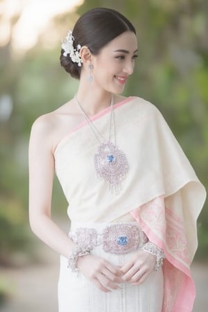 Portrait of A woman wearing a traditional Thai silk dress in soft White and pink embroidery, accessorized blue jewelry and a floral hairpiece. Her hair is neatly styled, and she is smiling gently in a bright and elegant setting, perfect for a formal occasion or ceremony.,Thai dress,soft focus,blurry foreground