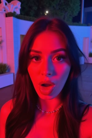 Generate a close-up, nighttime selfie in 8k resolution, featuring a young woman with a surprised and playful expression. The scene is illuminated by dramatic, colorful lighting, primarily a vibrant red hue from the left side, which casts strong highlights on the subject's face, and subtle cool blue lighting from the background. The contrast between the red and blue tones adds a lively, dynamic effect to the image.

The woman has long, dark, slightly tousled hair that frames her face. Her wide eyes and raised eyebrows give her an exaggerated, playful look of surprise. Her mouth is open, further enhancing the expressive, candid nature of the shot. She wears subtle makeup with winged eyeliner that accentuates her wide-eyed expression, and a soft, glossy lipstick that catches the light from the dramatic red glow. She is wearing a delicate necklace with small beads that shimmer faintly in the contrasting light.

In the background, the setting is outdoors at night, featuring a white textured wall with a few potted plants and a stylized design or signage partially visible. There are some indistinct buildings and greenery around, indicating an urban or event setting. The lighting in the background is subtle, with hints of blue that give depth and add a sense of nighttime ambiance without distracting from the subject.

The entire image has a spontaneous, lively vibe, emphasizing the woman’s playful, candid expression. The strong red and blue lighting contrast gives the scene an energetic and vibrant feel, making the subject's facial features and expression the focal point of the image.