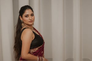 a medium-sized sexy indian woman stands in sidewise in front of a white curtain. She is dressed in a black sleeveless blouse with a maroon sari adorned with gold embroidery. Her hair is pulled back in a ponytail, and she is adorned with a pair of dangling earrings. Her bangles are gold, adding a touch of contrast to her dress. The backdrop is a stark white, creating a stark contrast to the woman's outfit.,photorealistic,Anupama, Warm Lighting,TamannaFlux, IMG0095.HEICl Viewing at camera in a  sensual way