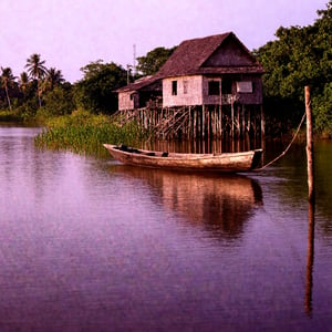 A realistic panoramic view of A traditional Malay wooden house stands gracefully by a calm riverside, surrounded by lush greenery. The house, raised on sturdy stilts, features intricate wooden carvings on its windows and doors, with a small verandah extending towards the river. Near the water's edge, a wooden sampan rests gently, tied to a wooden post. The scene is peaceful, with reflections of the house and trees rippling in the river, giving a sense of timeless rural life.real alamy, perfect anatomy, perfect image 