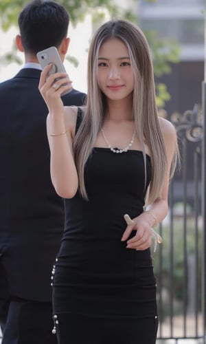 18 year old ,pale skin, a thai woman with long highlight white and darkbrown hair, wearing a slimfit sleeveless black dress adorned with white pearls, is smiling while holding a silver IPhone in her right hand. She is wearing a gold ring on her left hand, adding a touch of touch to her outfit. The background is blurred, suggesting a man in a black suit and dark hair is standing behind her.