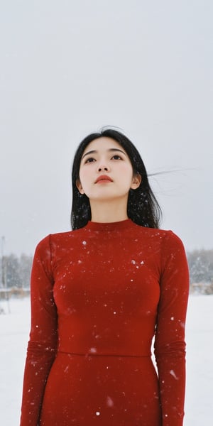 A young woman stands under a blanket of snowflakes gently falling from the gray winter sky. She wears a tight-fitting red dress, her long hair blowing in the gentle breeze as she gazes upwards, lost in thought. The camera frames her from the waist up, with the snowy landscape blurred behind her.,Film Photography