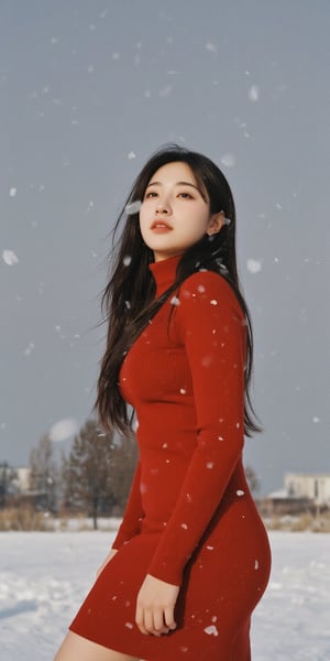 A young woman stands under a blanket of snowflakes gently falling from the gray winter sky. She wears a tight-fitting red dress, her long hair blowing in the gentle breeze as she gazes upwards, lost in thought. The camera frames her from the waist up, with the snowy landscape blurred behind her.,Film Photography