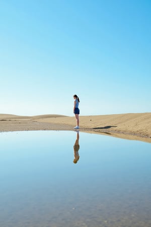 An extra long shot landscape photo showing a young  woman standing on a patch of sand, barely above the lack water. She has a ponytail, wears a blue tennis outfit, and She is looking at the horizon. We see her relfection in the water.  Background is a blue sky. The overall image shows a minimalistic and dreamini style.