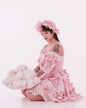 A full body shot of a woman, she's sitting down, she has split dyed hair (white and black), shes wearing a ruffled pastel pink dress on with tons of bows and ribbons, shes holding a giant white fluffy cloud in her hands, she looking down at her hands, she has on a light pink umbrella hat on, the background is white.