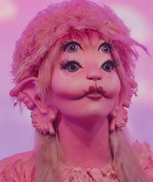 a vibrant pink haired girl with long, wavy blonde hair, wearing a pink fluffy hat, is adorned with a pink ribbon. She's dressed in a long-sleeved pink dress. Her eyes are wide open, adding a pop of color to the scene. The background is blurred, creating a stark contrast to the girl's outfit. The backdrop is a mix of pink and purple hues with little pink heart clouds, adding depth to the composition.