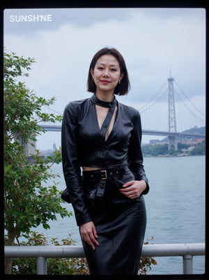 Captured on a digital screen, a woman stands in front of a body of water. She is dressed in a black leather jacket, a black skirt, and a black belt. Her hair is styled in a sleek bob, and her eyes are focused on the camera. The woman's hair is dark brown, and she is wearing a pair of black laces. The backdrop is a cloudy sky, and there is a bridge in the distance. To the left of the woman is a tree with green leaves, and the word "SUNSHINE" is visible in the top left corner of the screen. Below the screen, the words "Weekly Best Dressed" are written in white.