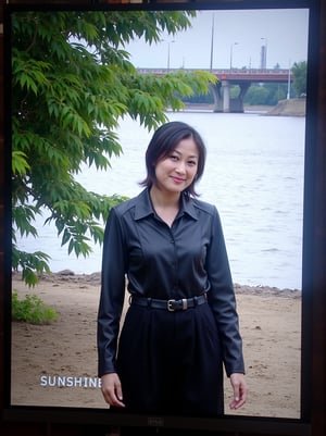 Captured on a digital screen, a woman stands in front of a body of water. She is dressed in a black leather jacket, a black skirt, and a black belt. Her hair is styled in a sleek bob, and her eyes are focused on the camera. The woman's hair is dark brown, and she is wearing a pair of black laces. The backdrop is a cloudy sky, and there is a bridge in the distance. To the left of the woman is a tree with green leaves, and the word "SUNSHINE" is visible in the top left corner of the screen. Below the screen, the words "Weekly Best Dressed" are written in white.
