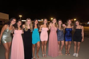 Group photo of several middle school girls all dressed up for their semiformal dance. All of the young girls are beautiful, each wearing a different beautiful dress that accentuates their developing figures. All of the girls are thin with a variety of hair colors and skin complexions. The photo is taken outside of the middle school gym at night using a high resolution Android phone. Each of the girls has a facial expression that shows their excitement for the night of dancing and frivolity. 