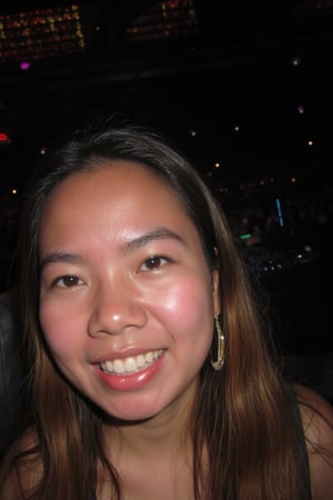 A close-up portrait captures an asian teenager's radiant smile at a lively party. Soft focus highlights their sparkling eyes and rosy cheeks. The dimly lit venue casts a warm glow on their face, illuminated by the flashing lights of the DJ booth in the background. A subtle blur effect suggests the chaos and energy of the celebration, while the subject's confident pose conveys excitement and belonging.