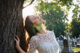 Photo.  Profile of a woman in a lace summer dress, She is looking upward with her eyes closed beside a tree.  Morning sun. Background is a cemetery