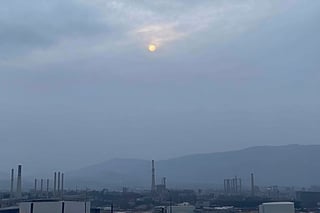 a dense layer of gray clouds illuminated in light rose color from above, a small golden sun, light gray mountains in the distance on the horizon among which power plants and industrial buildings can be seen, closer gray mountains on the horizon among which power plants and industrial buildings can be seen, black mountains on the horizon among which power plants and industrial buildings can be seen,