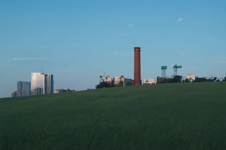 a huge hilly field covered with grass with partially clayey inclusions, green and brown boxes are visible very far away, also behind the hills narrow black skyscrapers with white windows with a yellowish tint are visible, green and brown boxes are visible far away, a dimly lit and dull black building with an orangish glow from sunset, you can see a small white bright advertisement with incomprehensible outlines, black skyscrapers with light yellow windows topped with metal structures standing right in the middle of the field reflect the light of the moon, in the distance you can see small two-story buildings with very small windows and a chimney, a little closer near the skyscrapers you can also see small two-story buildings buildings with very small windows and a chimney, not all skyscrapers are topped with metal structures, the sky on the horizon is from dark red to yellowish which sees off the departing sun, the moon is on the opposite side, the sky is soft blue topped by the moon on the opposite side of the sun, and a few stars, clouds light white very transparent barely visible,