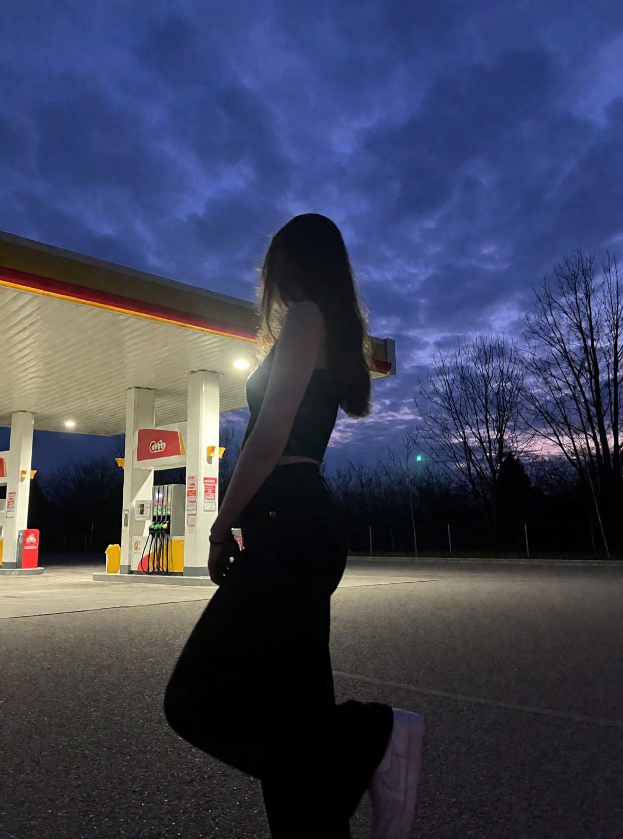 r3ealism,  Captured at dusk,  this low-light photograph features a silhouette of a person standing at a gas station by qwi...