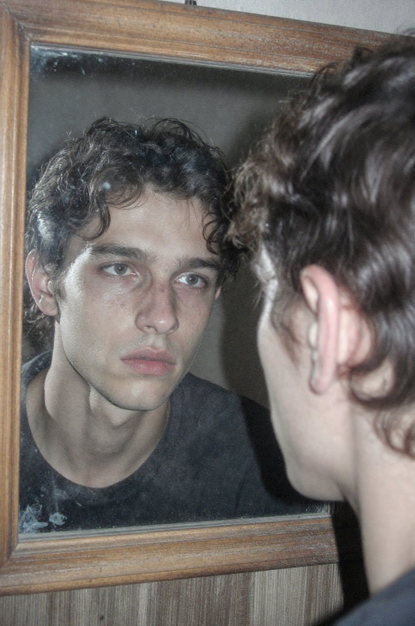 A gritty,  atmospheric over-the-shoulder portrait capturing the reflection of a young man with messy,  textured dark curly...
