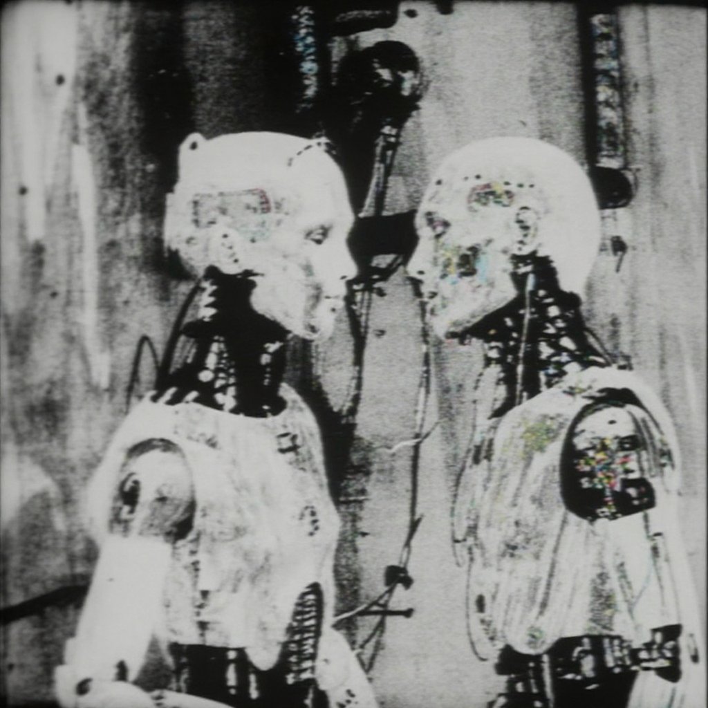 two humanoid androids standing chest-to-chest in a narrow industrial room; both have smooth white synthetic heads with min...
