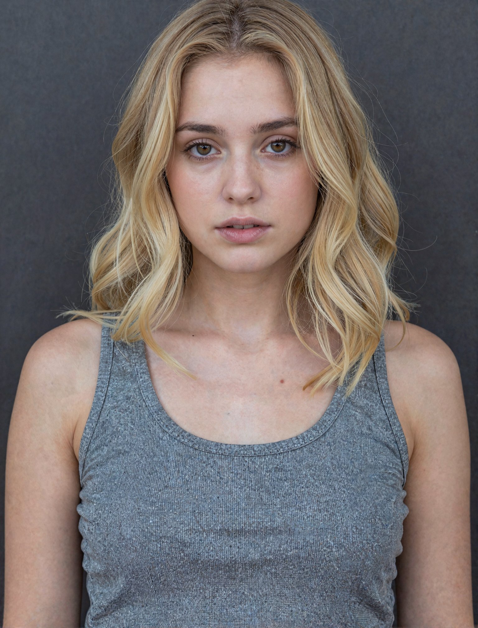 The image is a portrait of a young woman with shoulder-length blonde hair. She is wearing a gray tank top and is standing...