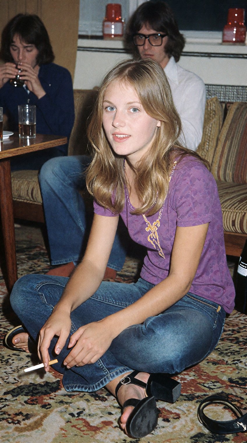 a woman with shoulder-length blonde hair sits cross-legged on a patterned carpet,  wearing a purple short-sleeved top,  da...