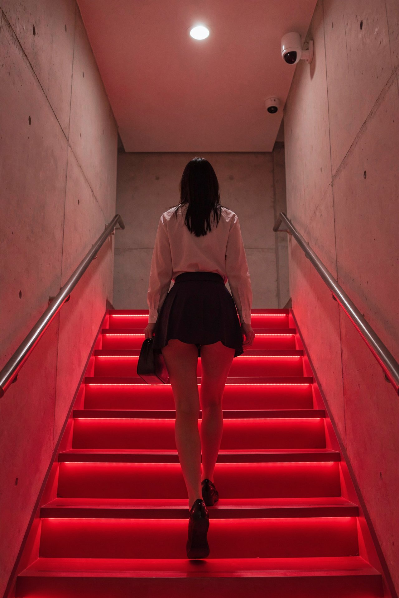 rear view of a person climbing a brightly red lit staircase. the individual has dark hair and is wearing a dark ultra mini...