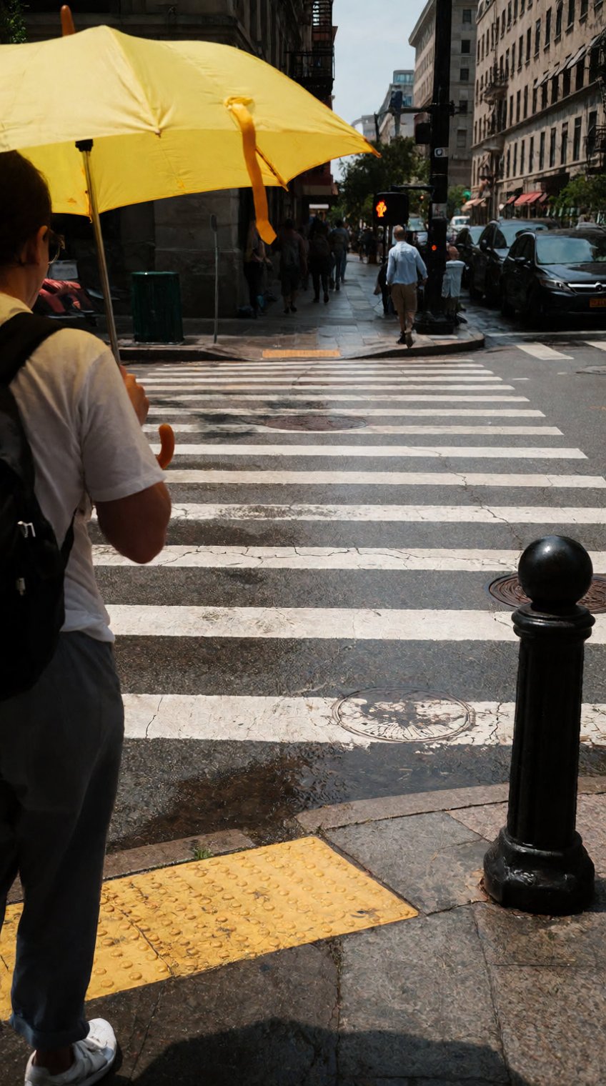 An eye-level candid photograph of a city crosswalk. In the foreground,  a person walks by holding a bright yellow umbrella...