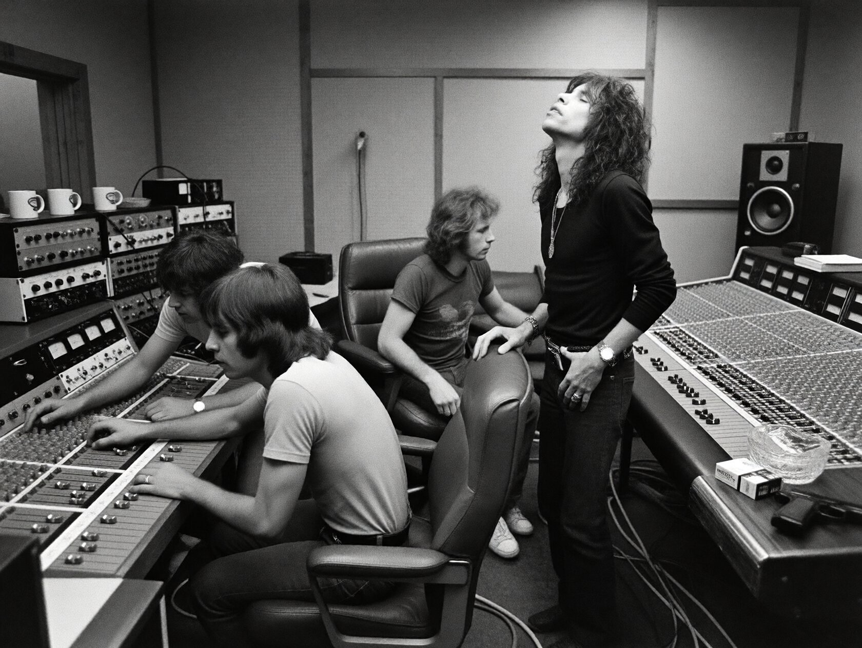 {"high_level_description":"A gritty,  high-contrast monochrome 1970s analog recording studio scene. Three men are focused...