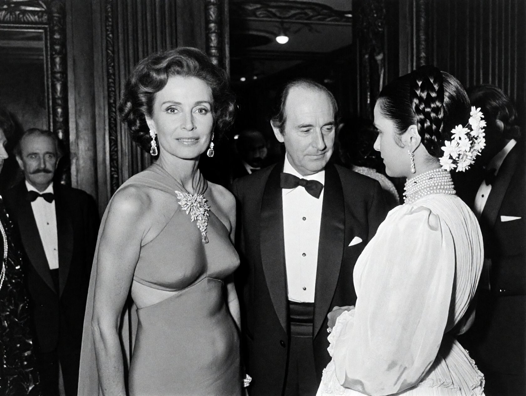 {"high_level_description":"A grain-heavy,  black and white candid photograph from a 1970s high-society gala. Three elegant...
