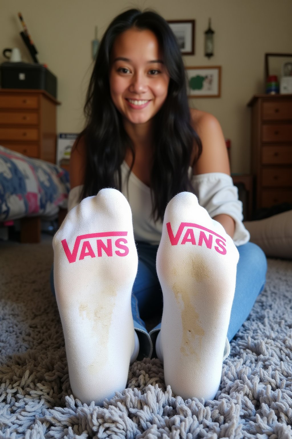 The image is a photograph of a young woman with a light skin tone and long, dark hair, posing in a cozy, cluttered bedroom. She is positioned on a plush, grey shaggy rug, with her feet prominently displayed in the foreground. She is wearing white socks with the word "VANS" printed in pink on the soles, which are slightly worn and dirty, in fact the suggestion of her footprint appears in the dirt. suggesting she has been wearing them for some time. She is smiling from ear to ear. 