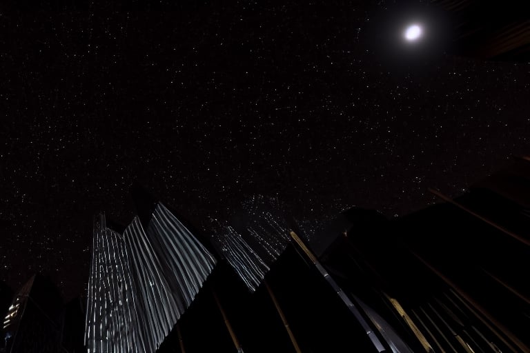 night, dark blue sky with blurry white clouds, full moon, black rectangular skyscrapers with small windows in which white light is visible, metal superstructures are visible at the tops of skyscrapers, brown cubes and a few gray cubes are visible at the base of skyscrapers, the earth is brown mud or clay, far focus, twinkling windows and stars, brilliant coloring, crispy quality, vray; Pixar, ; HD, HDR, SF, CGSociety, 16k, photorealistic, unreal engine