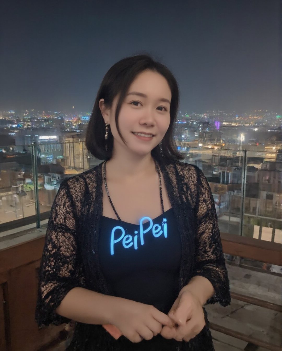 1girl, solo, (peipeiL, at the age of 20) with short dark hair, smiling, wearing an elegant black lace dress. She is adorned with a delicate necklace and matching earrings that shimmer under the city lights. Across her chest, glowing blue letters spell out 'PeiPei' in a light-up font, adding a unique and stylish touch. The backdrop is a stunning, enchanting Taipei nightscape. The sky is dark, with a few colorful city lights twinkling in the distance, reflecting the vibrant energy of the city. The scene is realistic, with a soft, warm glow highlighting PeipeiL's figure as she stands out against the alluring urban landscape. street,night,sky,realistic