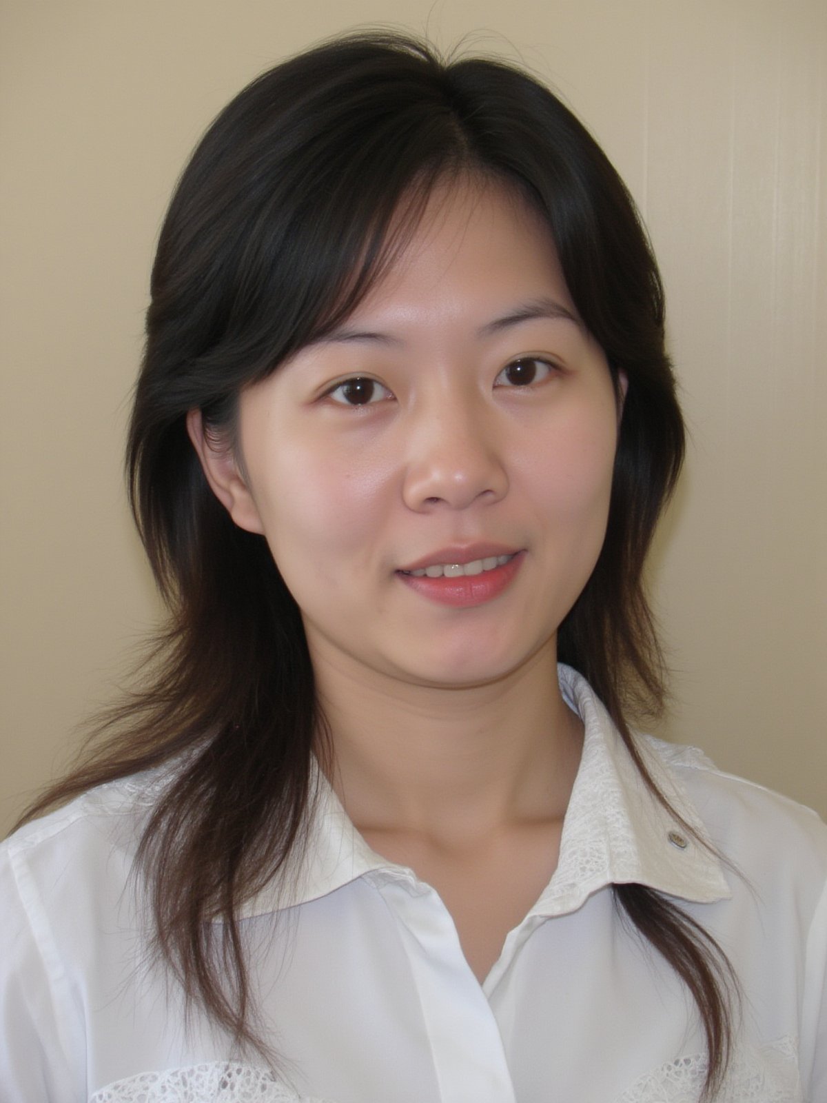 A close-up shot of a young Asian woman with long dark brown hair and brown eyes. She is wearing a white collared button down shirt with white embroidery on the collar. Her lips are a vibrant shade of pink. Her eyebrows are a light brown and her eyes are a darker shade of brown. She has a slight smile on her face. The backdrop is a light beige wall.