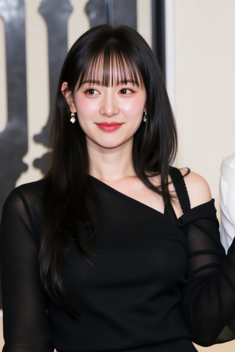 The image is a portrait of a young woman with long dark hair and bangs. She is wearing a black off-the-shoulder top with a high neckline and long sleeves. She has a pair of dangling earrings and is smiling at the camera. The background is blurred, but it appears to be an indoor setting with a white wall and a black and white patterned backdrop. The woman is posing for the camera with a slight smile on her face.