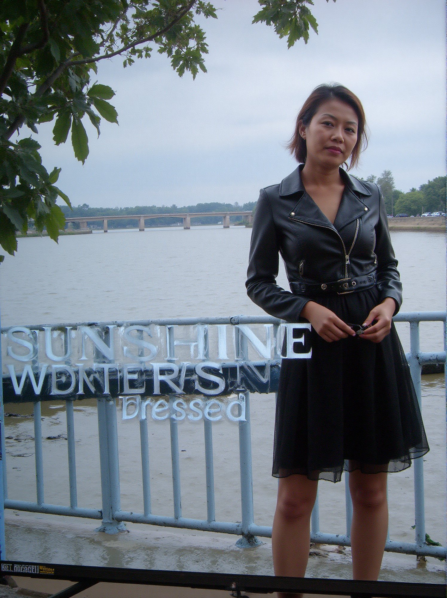 Captured on a digital screen, a woman stands in front of a body of water. She is dressed in a black leather jacket, a black skirt, and a black belt. Her hair is styled in a sleek bob, and her eyes are focused on the camera. The woman's hair is dark brown, and she is wearing a pair of black laces. The backdrop is a cloudy sky, and there is a bridge in the distance. To the left of the woman is a tree with green leaves, and the word "SUNSHINE" is visible in the top left corner of the screen. Below the screen, the words "Weekly Best Dressed" are written in white.