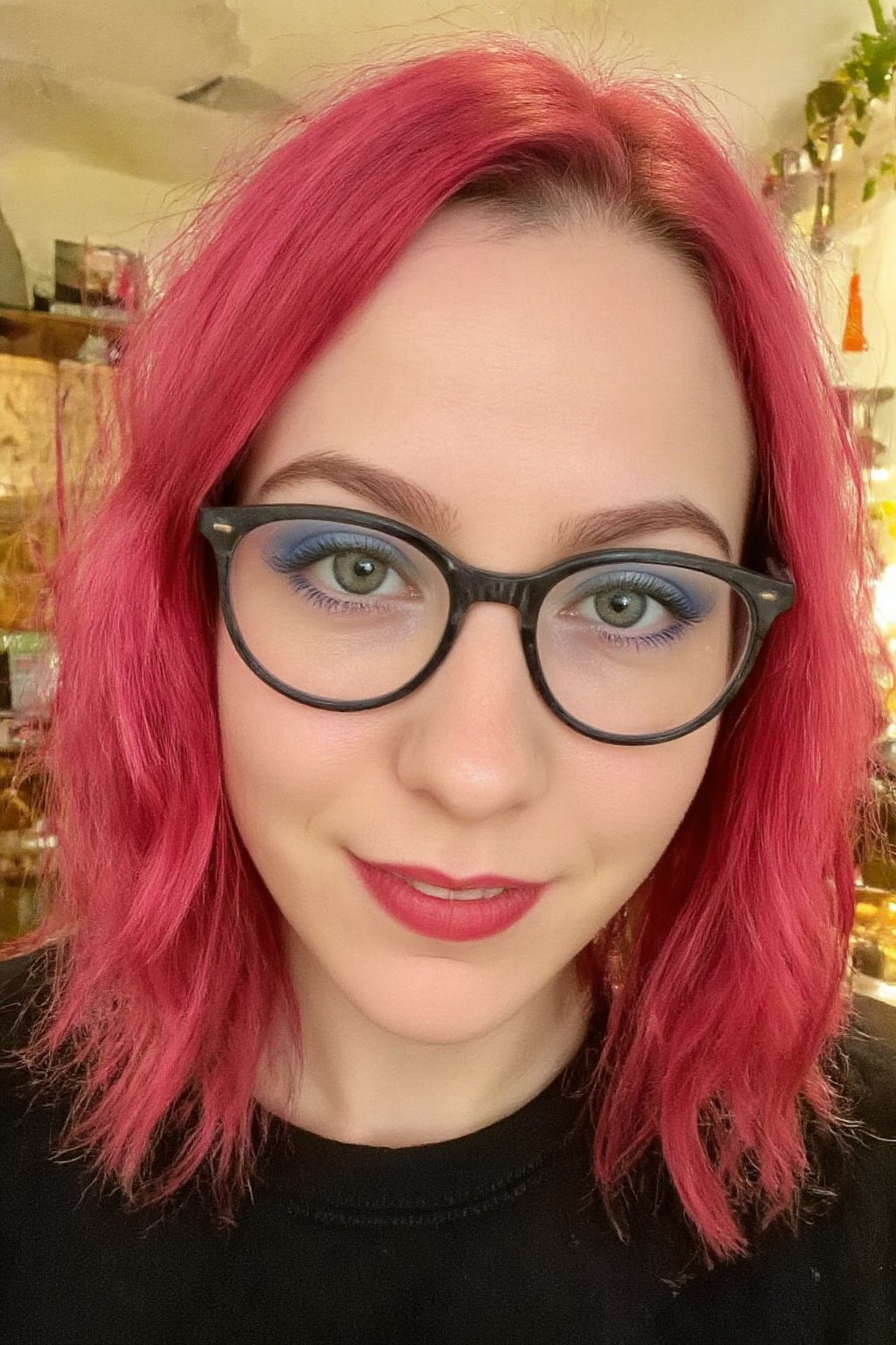 This is a close-up photograph of a 20yo western woman with striking features. She has vibrant, wavy pink hair cascading past her shoulders and wears black-rimmed glasses. Her skin is fair. Her eyes are green with bold blue eyeshadow, and she has a bright red lipstick on her full lips, which are slightly parted in a cheerful smile. She is wearing a black top. The background is slightly blurred but appears to be indoors, possibly a living room with warm lighting, wooden furniture, and some hanging decorations. The overall atmosphere is casual and vibrant, emphasizing the subject's colorful and expressive appearance.