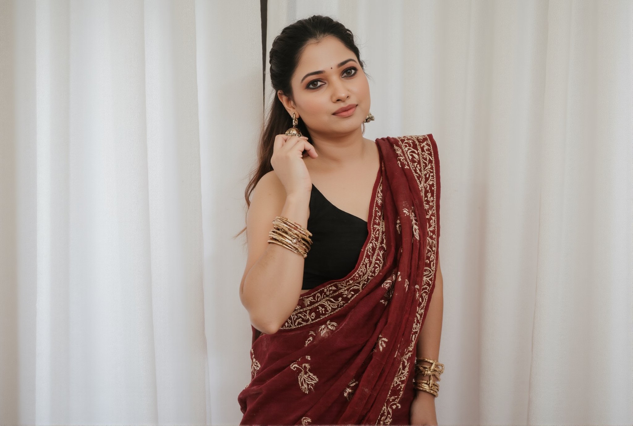 a medium-sized sexy indian woman stands in sidewise in front of a white curtain. She is dressed in a black sleeveless blouse with a maroon sari adorned with gold embroidery. Her hair is pulled back in a ponytail, and she is adorned with a pair of dangling earrings. Her bangles are gold, adding a touch of contrast to her dress. The backdrop is a stark white, creating a stark contrast to the woman's outfit.,photorealistic,Anupama, Warm Lighting,TamannaFlux, IMG0095.HEICl Viewing at camera in a  sensual way