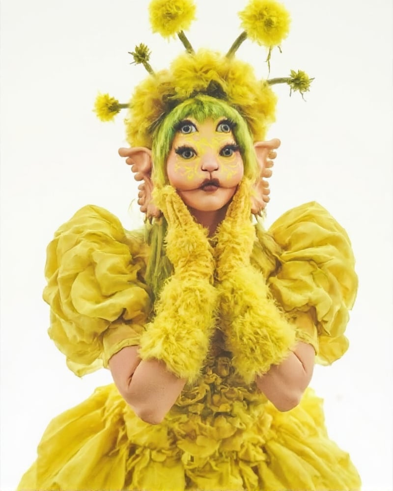 Close-up shot of a whimsical woman dressed in a bright yellow puffy dress, with multiple vibrant yellow flowers sprouting from her hair. Her hands are clad in matching yellow fluffy gloves, while her feet wear oversized yellow boots. Against a crisp white background, she stands out like a ray of sunshine, her sunny attire and accessories radiating joy and playfulness.