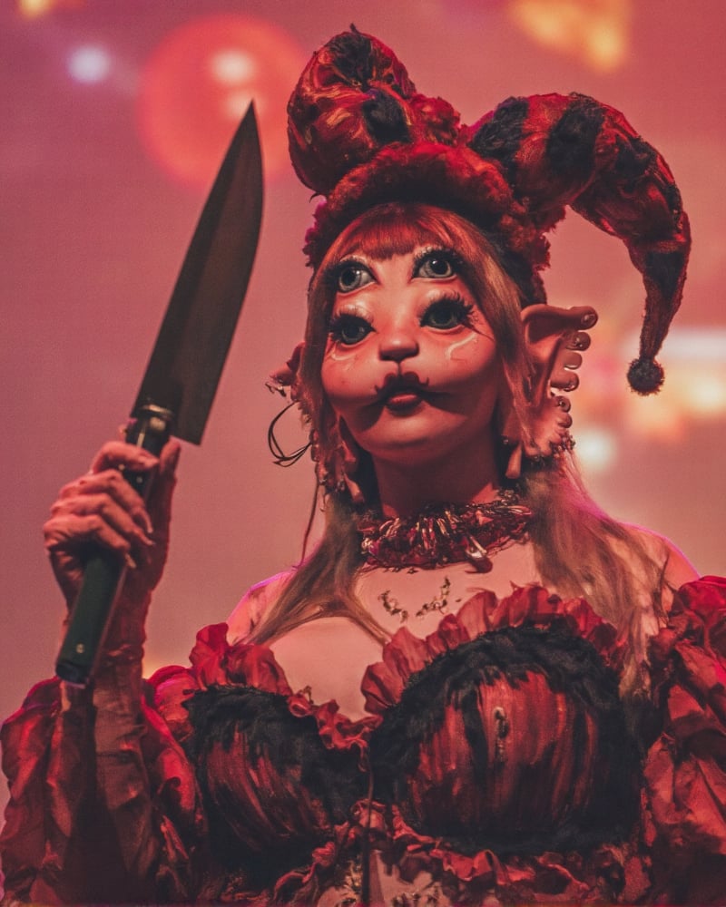 A stunning redhead stands confidently in the spotlight of a vibrant circus backdrop, her fiery locks contrasting with the bold red and black jester hat perched atop her head. A striking black and red clown suit wraps around her lithe physique, its colors echoed by the balloons floating above. In one hand, she grips a massive knife, its gleam catching the twinkling lights of the circus, as if ready to take on the whimsical world around her.