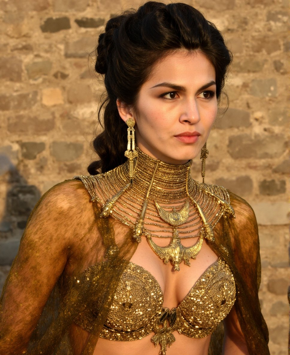 This is a high-resolution photograph of a woman of Middle Eastern or Mediterranean descent, likely in her mid-20s, with a fair to medium skin tone and dark, wavy hair pulled back into an elegant updo. She has a serious, contemplative expression, with her lips slightly parted and her eyes gazing off to the side. Her attire is elaborate and regal, consisting of an ornate golden breastplate adorned with intricate patterns and sequins that shimmer under the light. The breastplate is supported by a matching golden collar that extends up to her neck and shoulders, featuring delicate, interlocking designs. She also wears a long, sleeveless cape made of the same golden fabric, draped over her shoulders, which adds a touch of grandeur to her ensemble. The background is a rustic stone wall, suggesting a medieval or ancient setting, with rough, textured stones providing a backdrop that contrasts with her luxurious attire. The lighting is warm and soft, casting gentle shadows that enhance the richness of her attire and the texture of the stone wall. The overall atmosphere of the image is one of historical grandeur and nobility... <lora:Elodie_Yung_Flux_V1:0.96>