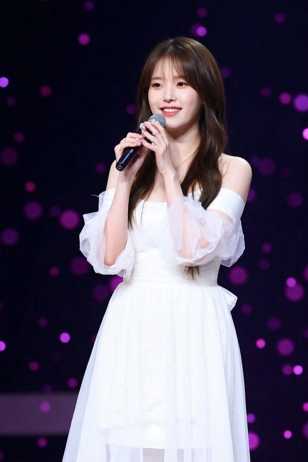 (medium full shot), beautiful young korean girl performing on stage, bathed in bright spotlight. She is wearing a white, off-shoulder dress with flowing, layered fabric, giving her an ethereal and elegant appearance. Her long hair cascades over her shoulders, and she holds a microphone close to her mouth, seemingly in the midst of a song. The background is dark with a bokeh effect of small pink and purple lights, suggesting a large audience or a concert setting. , <lora:flux_realism_lora:1>,  <lora:makinaflux_iu_v1.0:1>