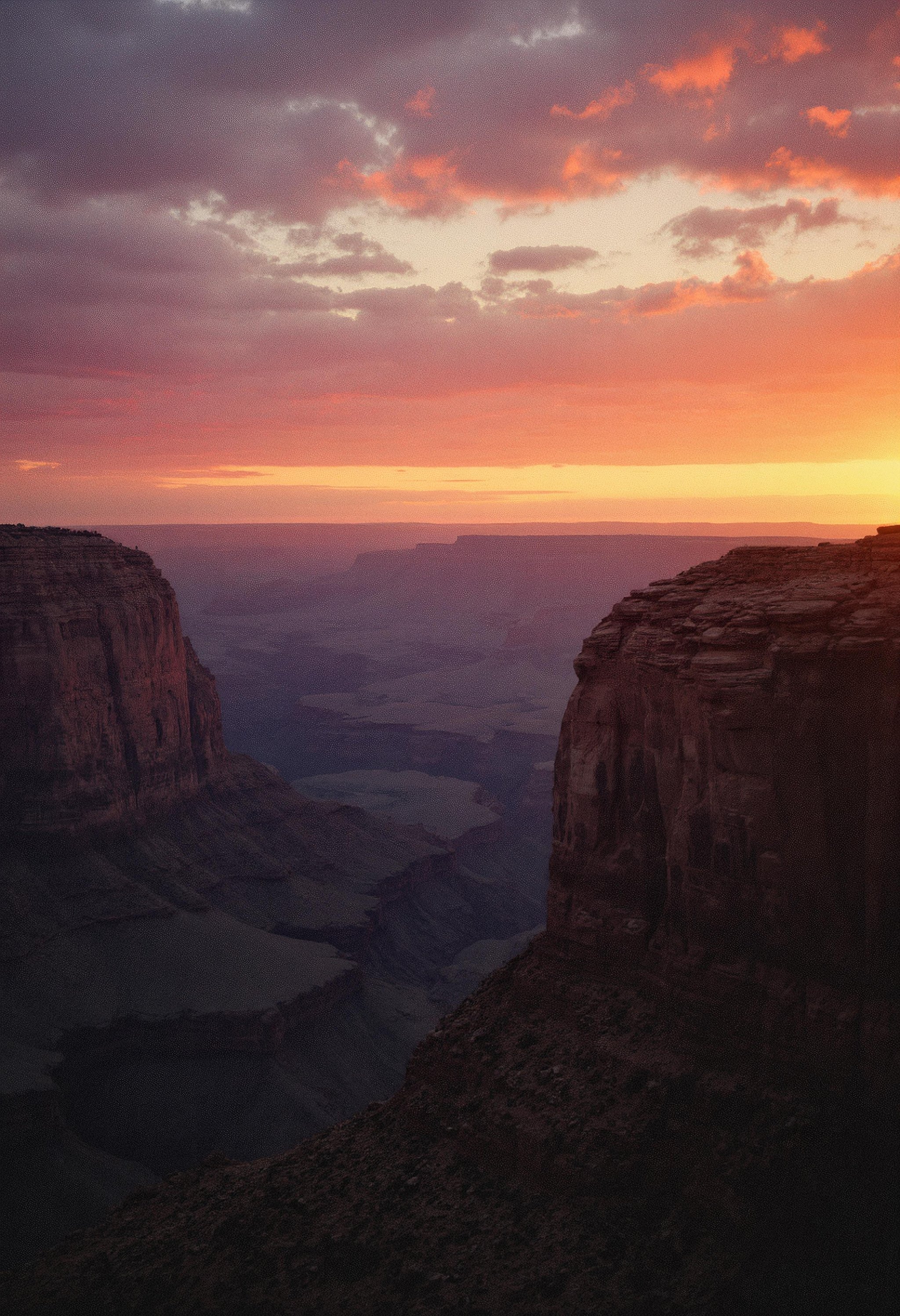 A wide landscape shot of a canyon, side-lit by the setting sun, casting dramatic shadows and highlighting the rugged textures of the rock formations. The sky is a stunning gradient of colors, transitioning from warm golds and oranges to deep purples and blues. The atmospheric depth creates a sense of vastness and dimension, with distant layers of the canyon fading into the horizon. The lighting emphasizes the intricate details of the canyon walls and the rich, vibrant hues of the sky. Grainy, grain, film grain, candid, high contrast, film burn, analog texture, partial silhouette, Chiaroscuro, off-centered, asymmetrical, highlight bloom, filmic glow, blown-out highlights, moody, herbstphoto, herbst photo. <lora:realism_lora:1.0> <lora:Herbst_Photo_Extra_Texture-0000020:2.0><lora:Herbst Photo Lora Test_00484_:0>